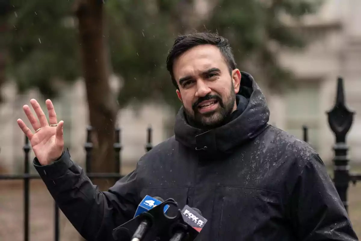 Hombre con barba y chaqueta oscura hablando frente a micrófonos al aire libre en un día lluvioso Hombre con barba y chaqueta oscura hablando frente a micrófonos al aire libre en un día lluvioso