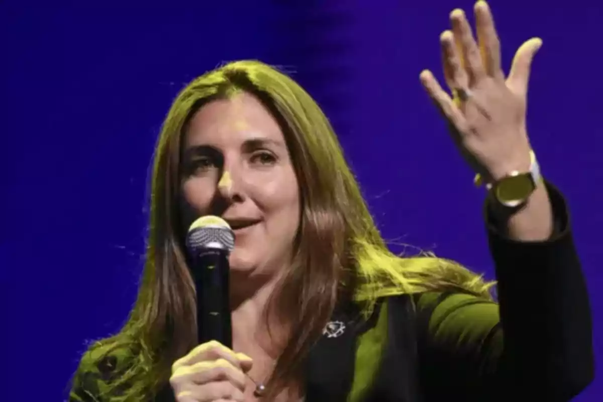 Long-haired woman speaking into a microphone with a blue background and raising one hand Long-haired woman speaking into a microphone with a blue background and raising one hand