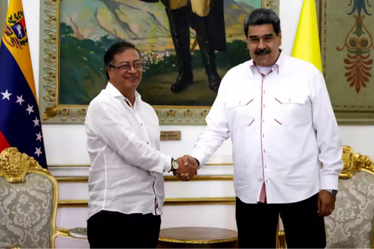 Two men dressed in white jerseys shake hands and smile in an elegant room with flags and a painting in the background.