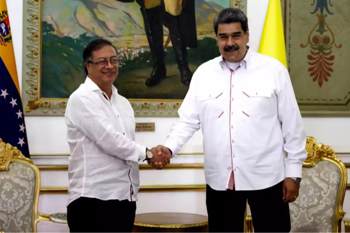 Two men standing shake hands and smile in an elegant room with golden chairs and a painting in the background.