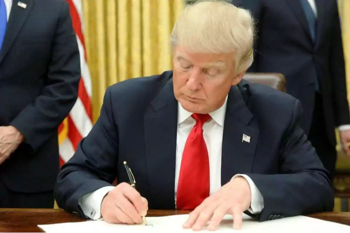 Blond-haired man in a dark suit and red tie signing a document at a desk