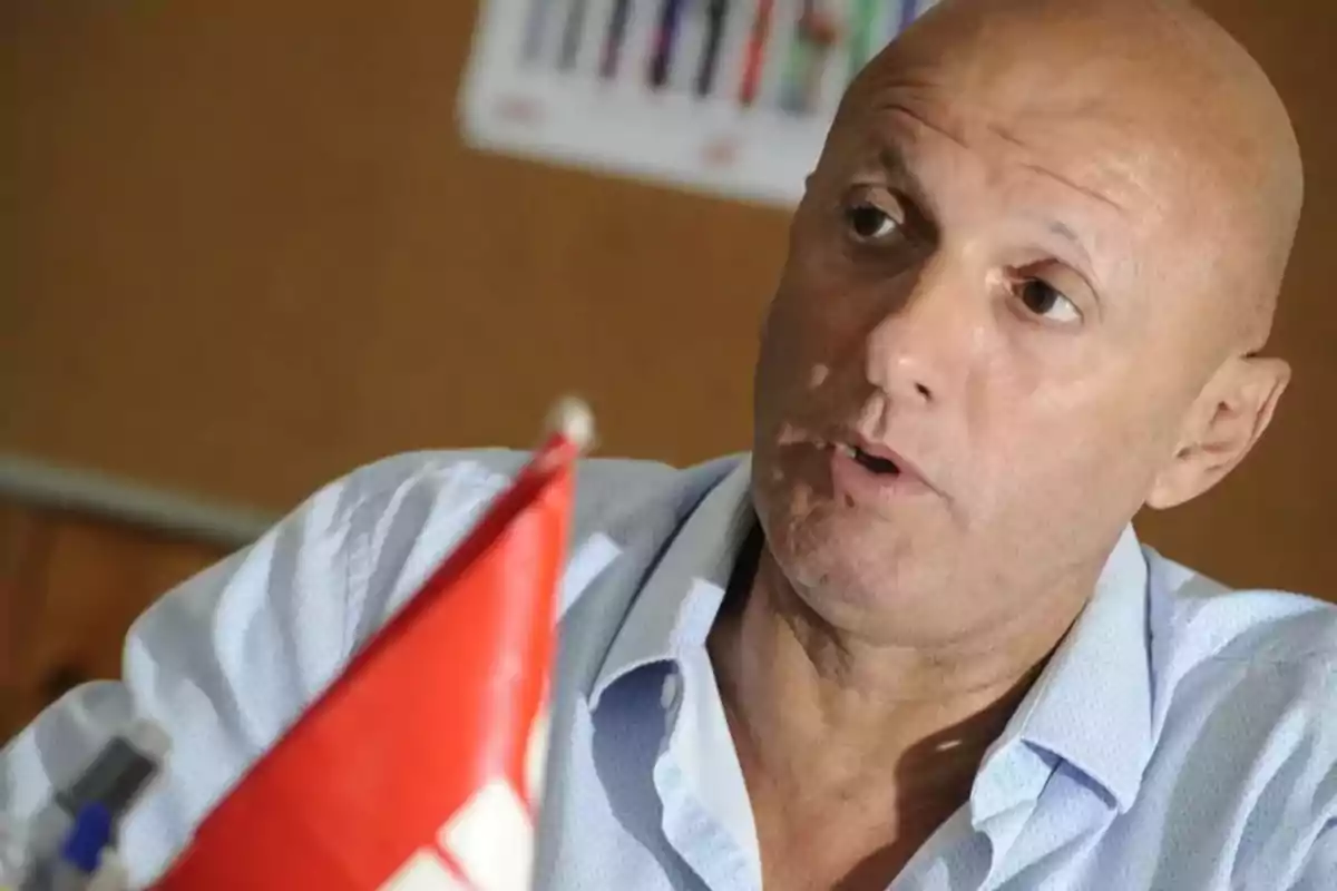 Hombre calvo con camisa clara hablando, con una bandera roja y blanca en primer plano y un fondo desenfocado. Hombre calvo con camisa clara hablando, con una bandera roja y blanca en primer plano y un fondo desenfocado.