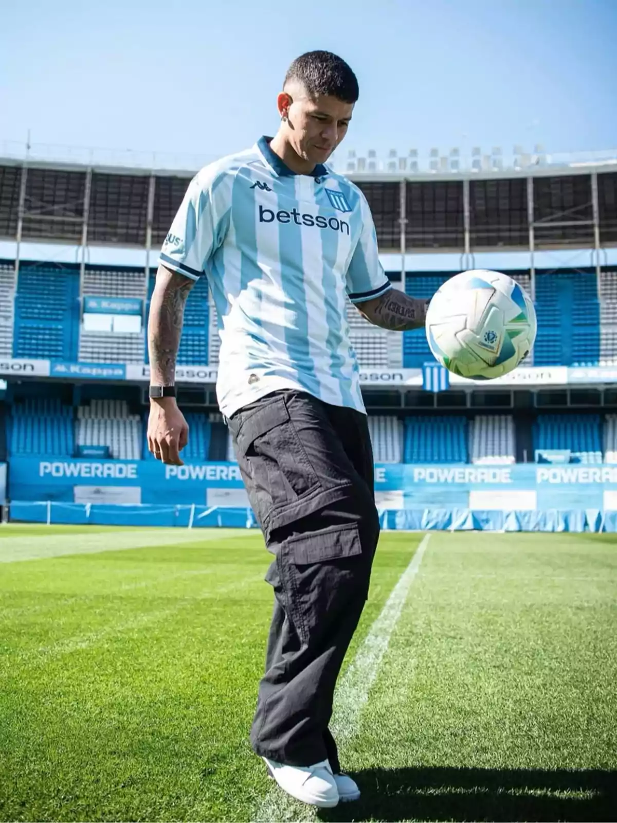 Un futbolista con la camiseta de Racing Club domina un balón en el césped de un estadio vacío en un día soleado