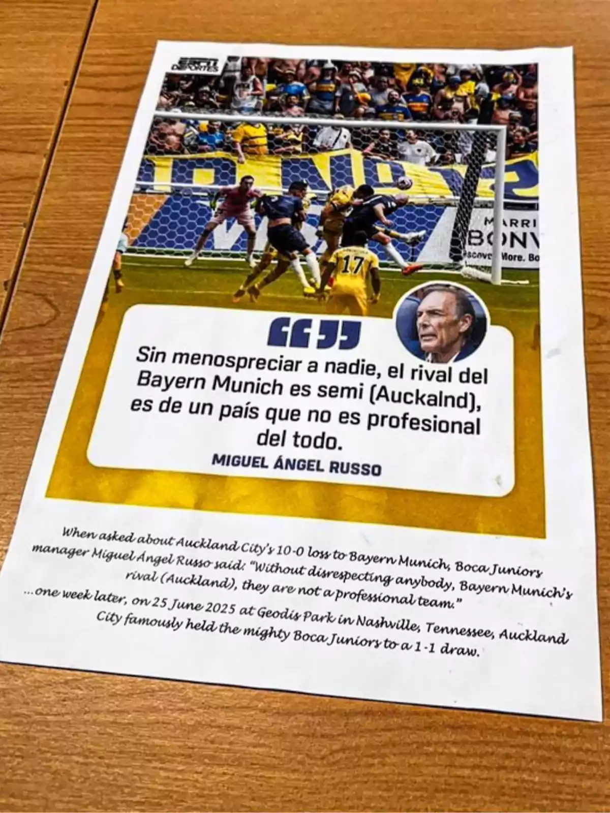 Printed sheet on a wooden table showing an image of a soccer match with players in the box and a quote from Miguel Ángel Russo about the Auckland City team, accompanied by a text in English that explains the context of the statement and the result of a subsequent match. Printed sheet on a wooden table showing an image of a soccer match with players in the box and a quote from Miguel Ángel Russo about the Auckland City team, accompanied by a text in English that explains the context of the statement and the result of a subsequent match.