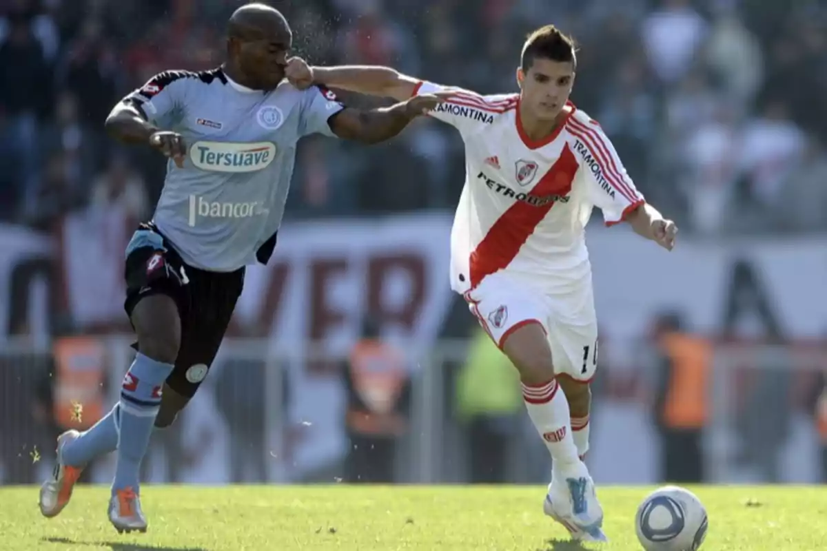 Dos futbolistas disputan el balón durante un partido uno con uniforme celeste y el otro con uniforme blanco con una banda roja Dos futbolistas disputan el balón durante un partido uno con uniforme celeste y el otro con uniforme blanco con una banda roja
