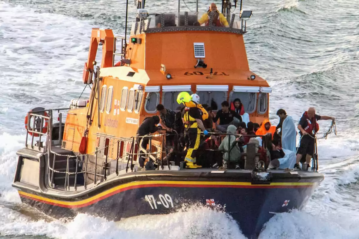 Un barco de rescate naranja navega por el mar con varias personas a bordo, algunas cubiertas con mantas y otras usando chalecos salvavidas, mientras miembros de la tripulación asisten a los pasajeros Un barco de rescate naranja navega por el mar con varias personas a bordo, algunas cubiertas con mantas y otras usando chalecos salvavidas, mientras miembros de la tripulación asisten a los pasajeros