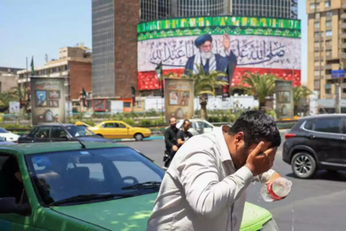 A man cools himself with water on a busy street while in the background a large billboard with the image of a religious leader can be seen and several cars are driving by.