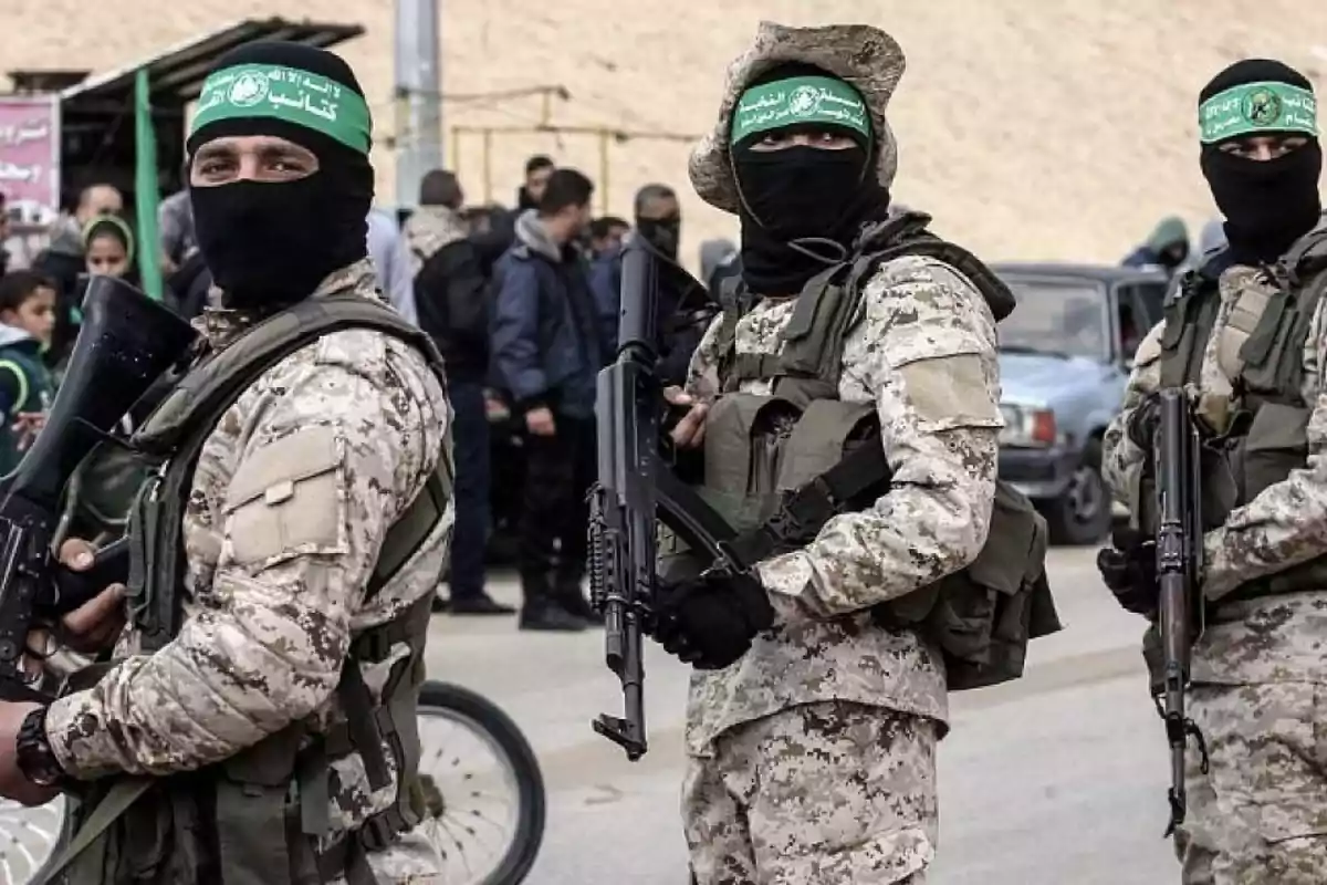 Three people in military uniforms and balaclavas are carrying weapons while standing on a street with more people in the background. Three people in military uniforms and balaclavas are carrying weapons while standing on a street with more people in the background.
