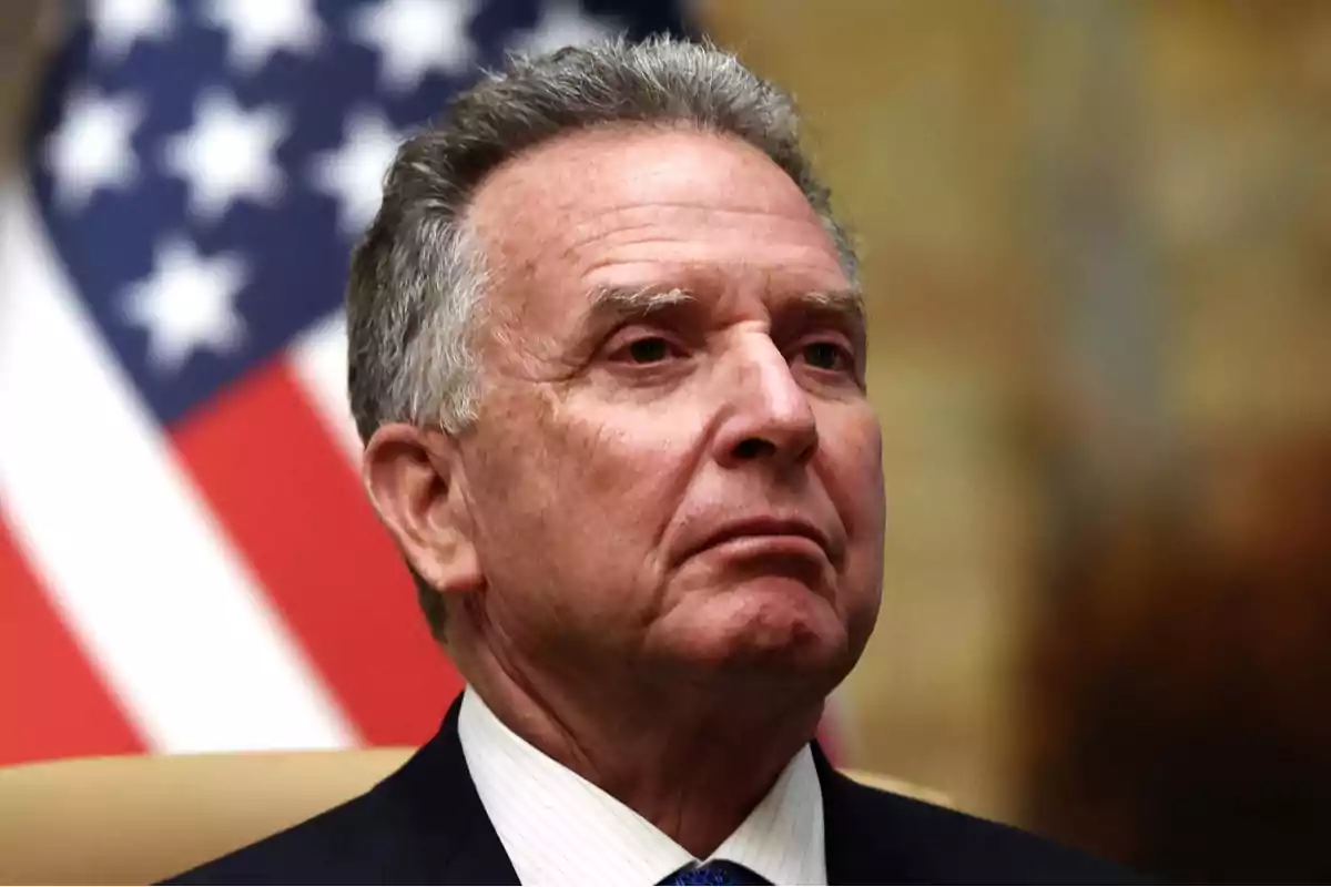 Hombre de cabello canoso con expresión seria y bandera de Estados Unidos desenfocada al fondo