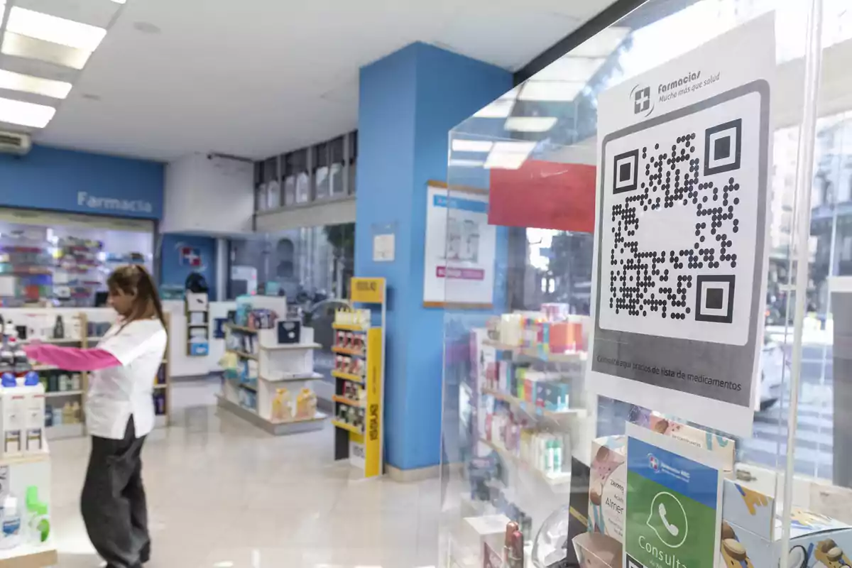 Interior de una farmacia con una empleada organizando productos y un código QR pegado en una mampara de vidrio Interior de una farmacia con una empleada organizando productos y un código QR pegado en una mampara de vidrio