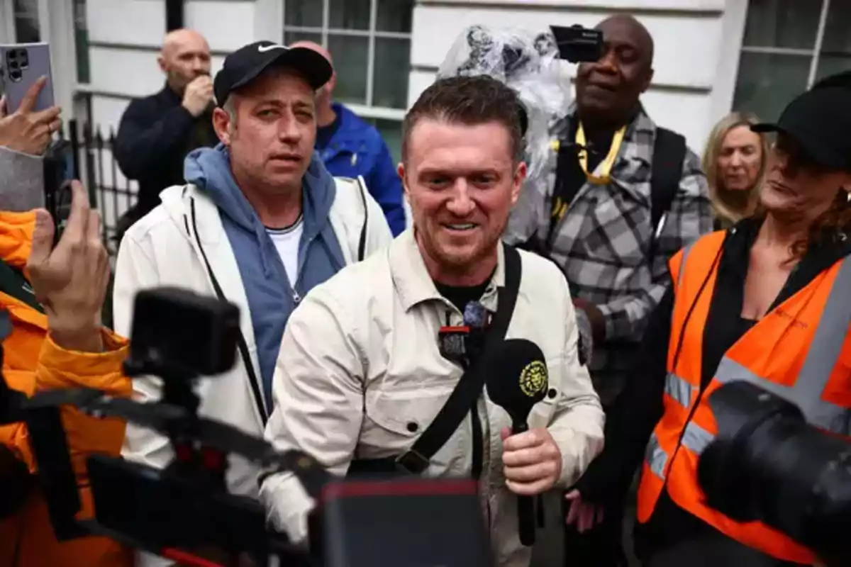 A smiling man holds a microphone surrounded by several people and cameras on a street. A smiling man holds a microphone surrounded by several people and cameras on a street.