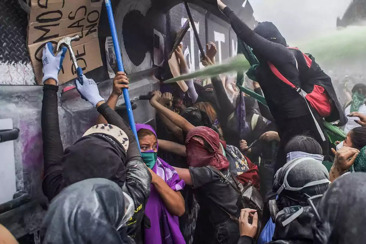 Group of women protesters with scarves and covered faces confront a metal barrier as a green substance is sprayed toward them. Group of women protesters with scarves and covered faces confront a metal barrier as a green substance is sprayed toward them.