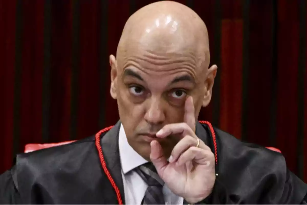 Hombre calvo con toga negra y corbata gris levantando el dedo índice frente a su rostro con expresión seria