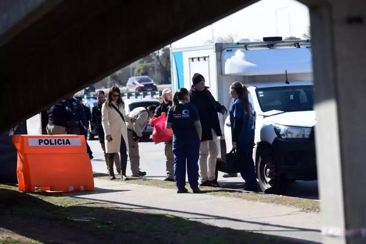 Varias personas, entre ellas policías y personal forense, se encuentran reunidas junto a una camioneta blanca y un cartel naranja de policía en una calle, bajo un puente. Varias personas, entre ellas policías y personal forense, se encuentran reunidas junto a una camioneta blanca y un cartel naranja de policía en una calle, bajo un puente.