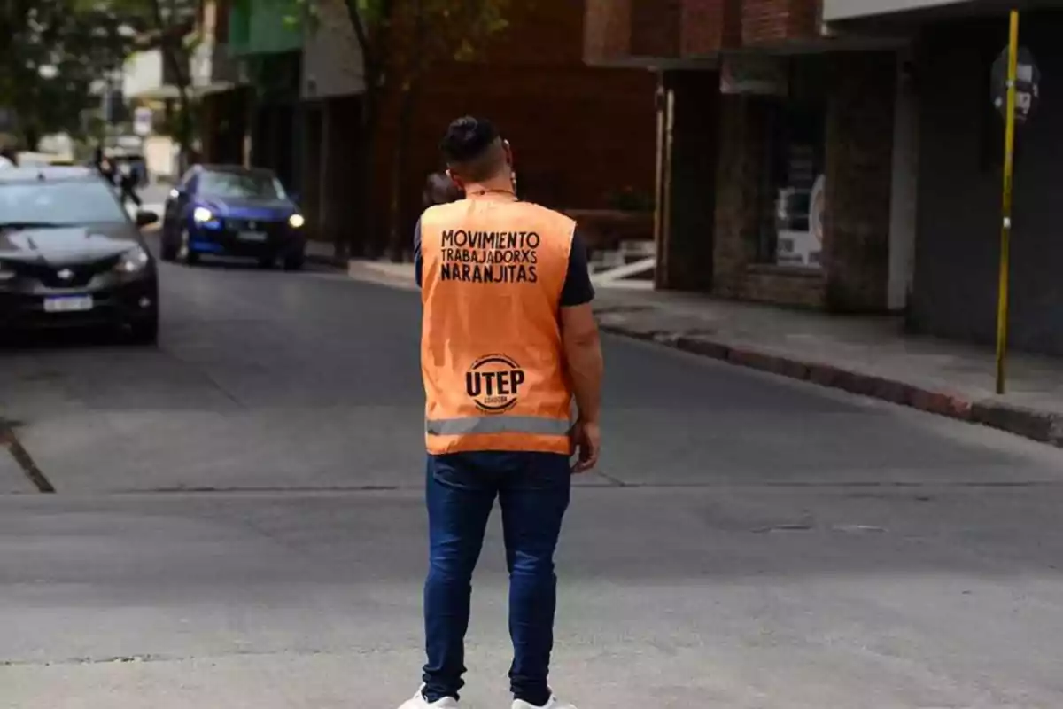 Person facing away wearing an orange vest that says Movimiento Trabajadorxs Naranjitas UTEP standing on an urban street