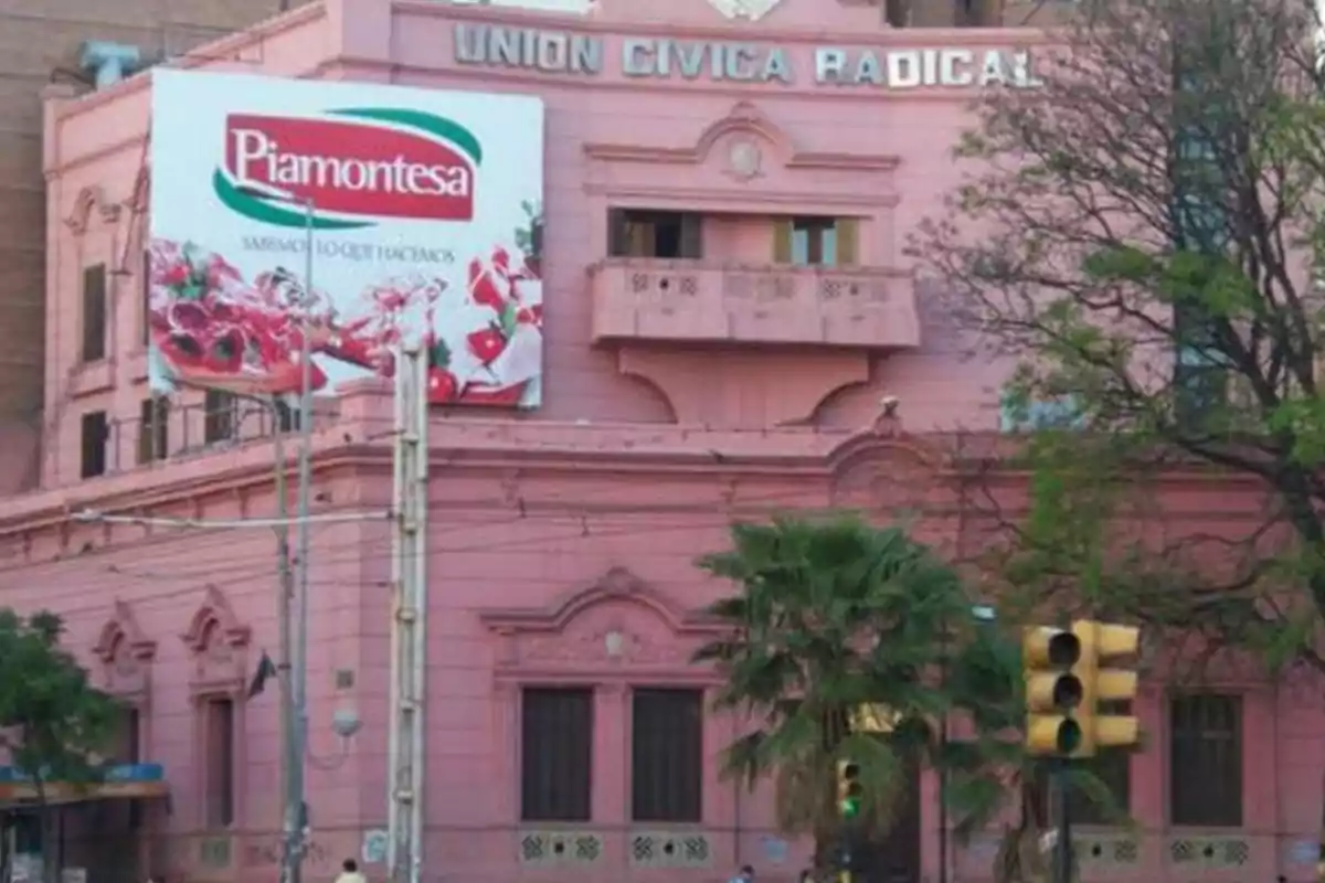 Old pink building with a Piamontesa billboard and a traffic light in the foreground surrounded by trees