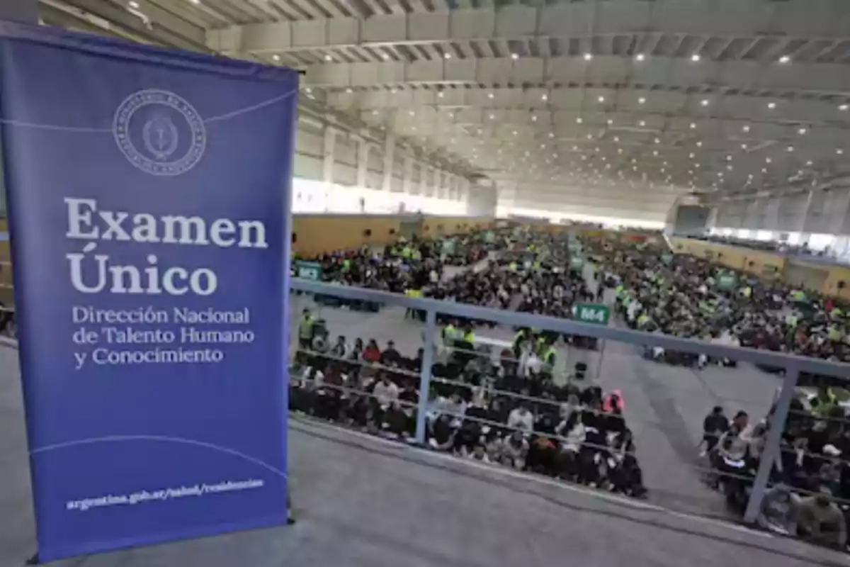 Gran salón con muchas personas sentadas rindiendo un examen y un cartel azul que dice Examen Único Dirección Nacional de Talento Humano y Conocimiento Gran salón con muchas personas sentadas rindiendo un examen y un cartel azul que dice Examen Único Dirección Nacional de Talento Humano y Conocimiento