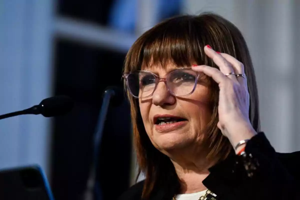 Mujer de cabello castaño y gafas hablando frente a un micrófono mientras se ajusta las gafas