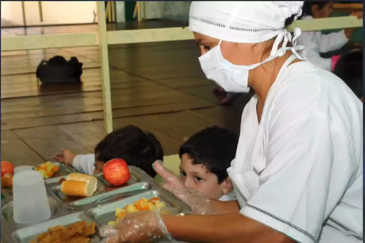 Mujer con uniforme y cubrebocas sirviendo comida a niños pequeños en una bandeja en un comedor escolar