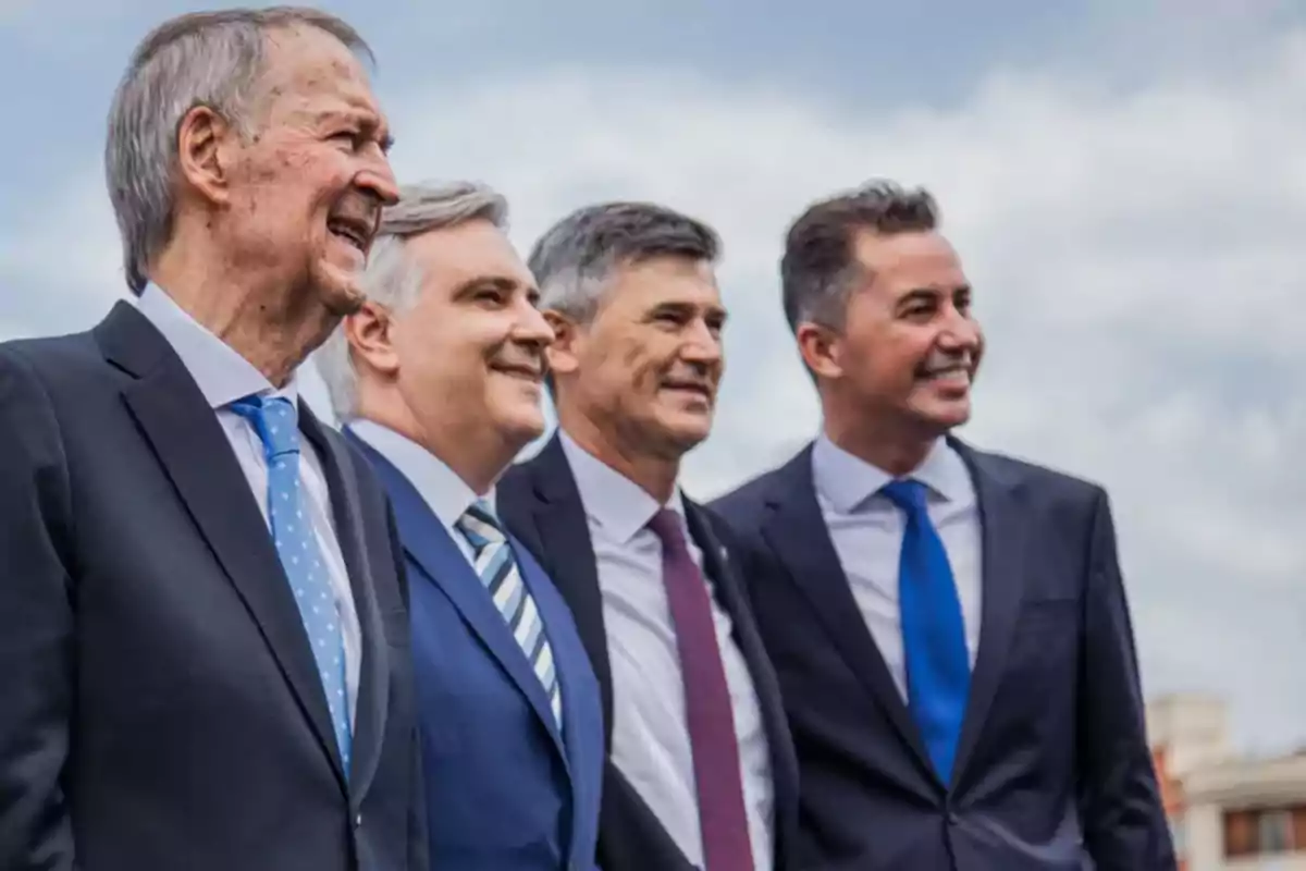 Cuatro hombres vestidos con traje y corbata posan sonrientes al aire libre bajo un cielo parcialmente nublado.