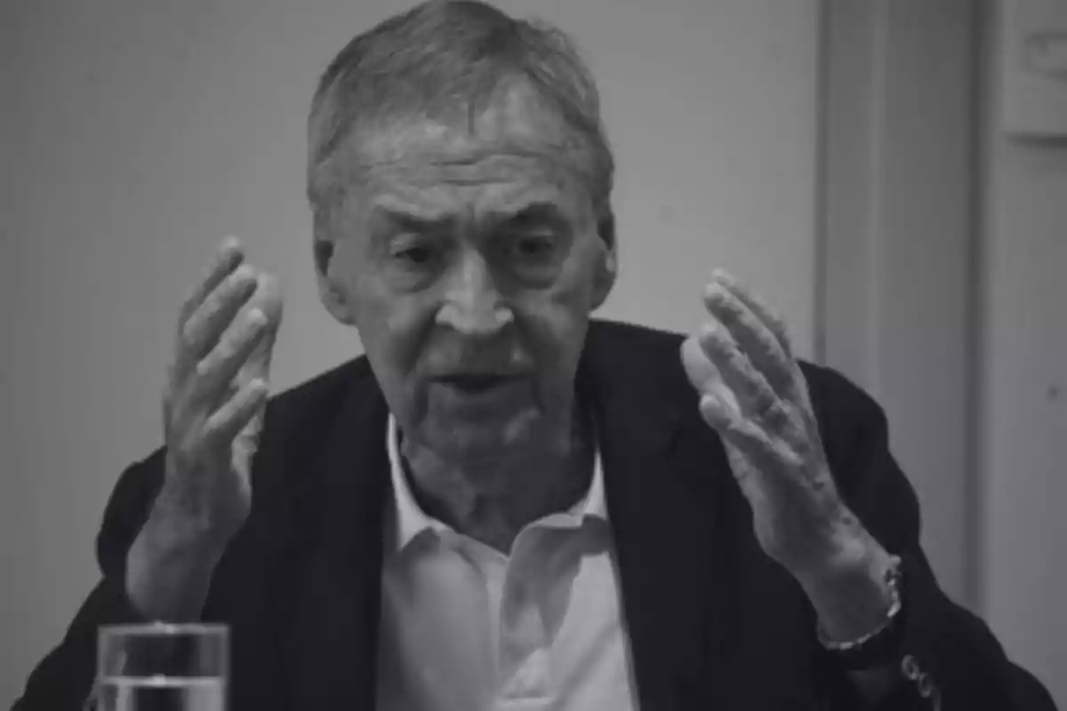 Older man with gray hair gesturing with both hands while speaking in a well-lit room