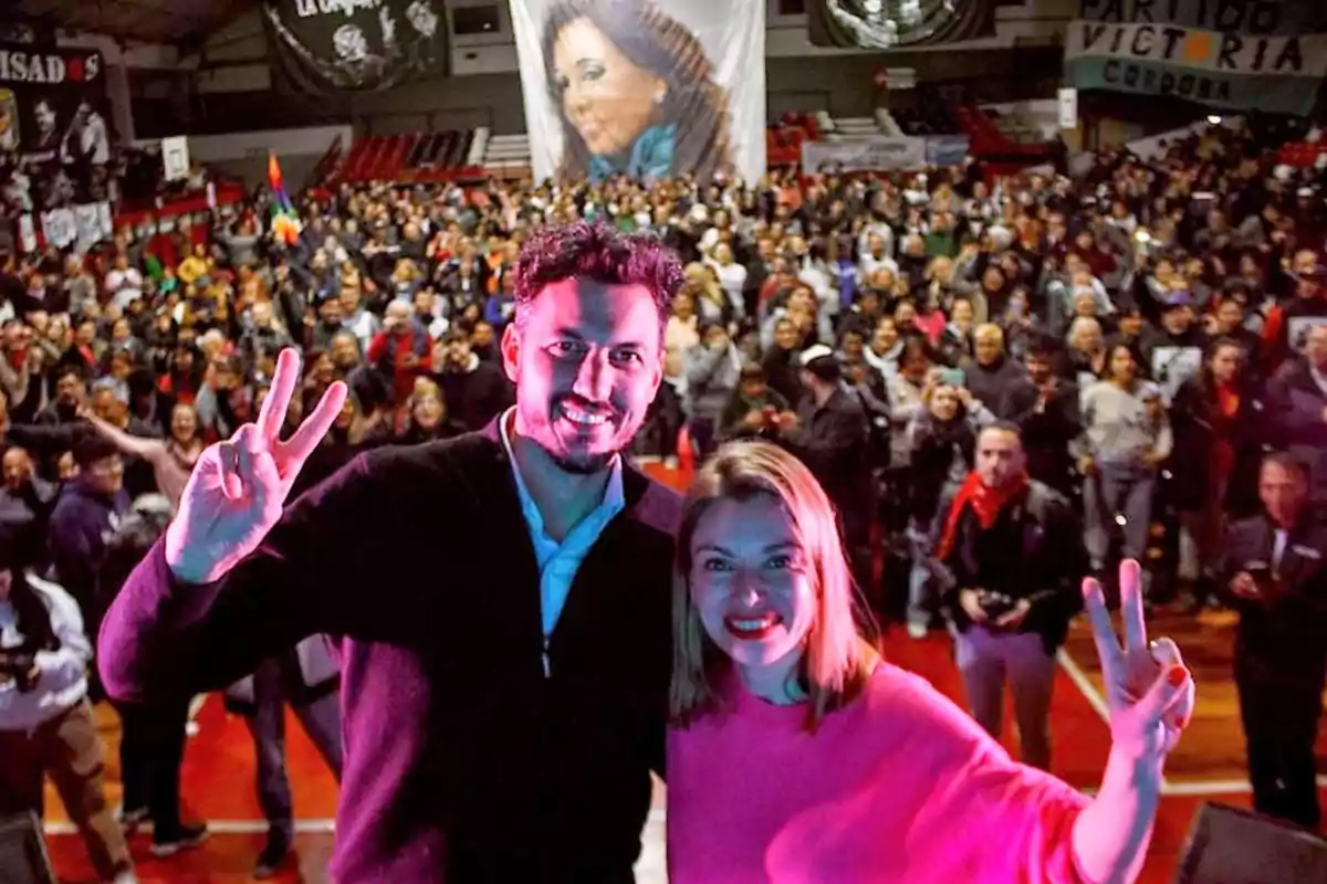 Dos personas sonríen y hacen la señal de la victoria con los dedos mientras posan para una selfie frente a una multitud en un estadio cubierto