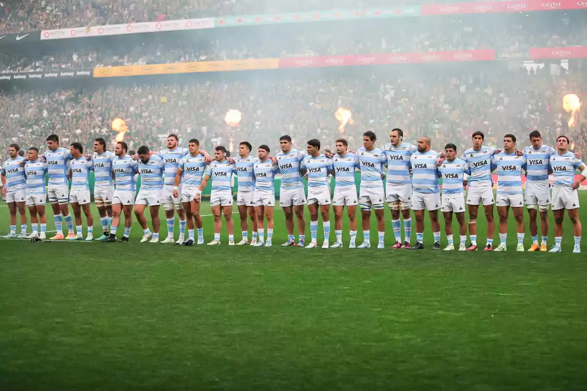 Jugadores de rugby con uniforme celeste y blanco alineados en el campo antes de un partido con público y fuegos artificiales de fondo Jugadores de rugby con uniforme celeste y blanco alineados en el campo antes de un partido con público y fuegos artificiales de fondo