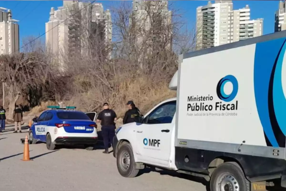 Several police officers and official vehicles from the Public Prosecutor's Office and the police are on a dirt road near tall buildings and dry trees.