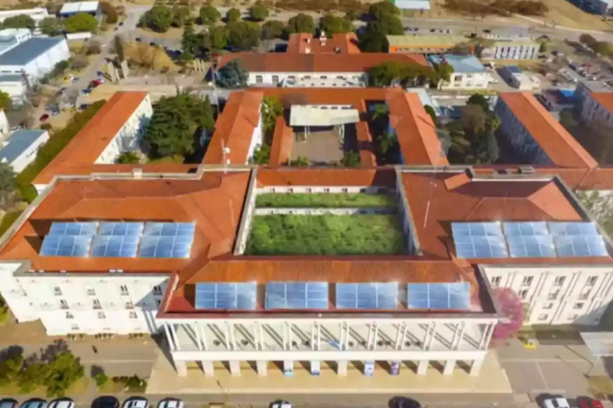 Vista aérea de un edificio grande con techo rojo y paneles solares, rodeado de áreas verdes y estacionamiento al frente