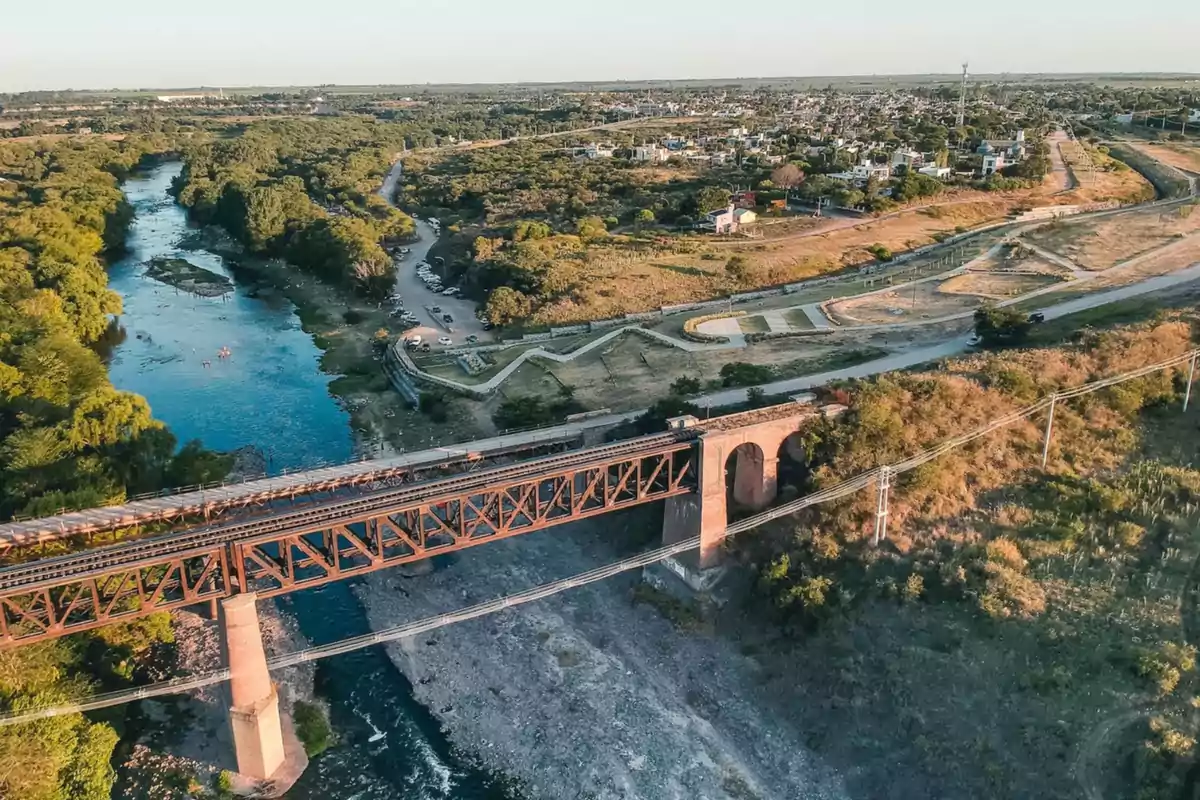 Vista aérea de un puente ferroviario antiguo sobre un río rodeado de vegetación y una ciudad al fondo