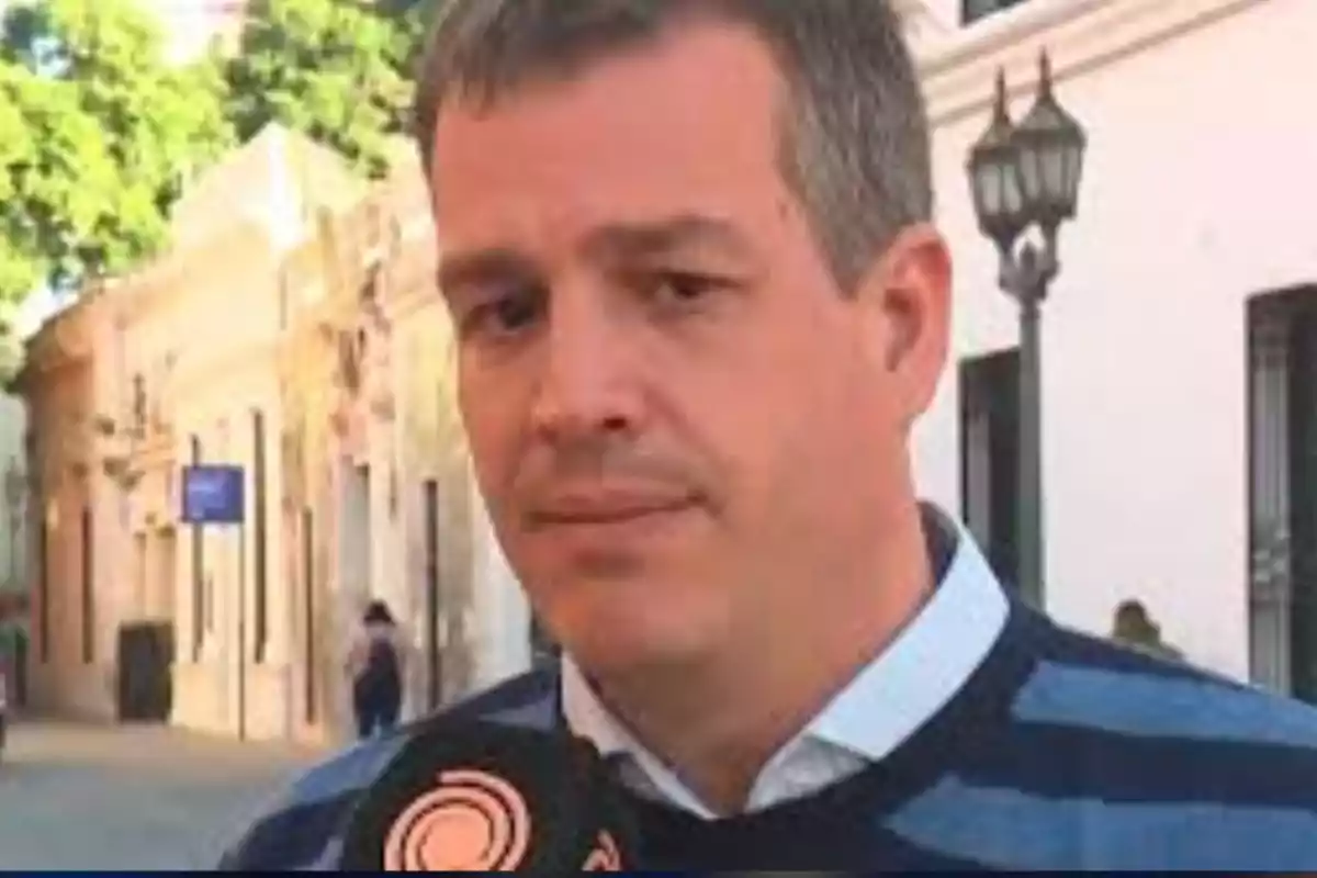 Hombre de cabello corto y canoso con suéter de rayas azules hablando frente a un micrófono en una calle con edificios antiguos y árboles al fondo