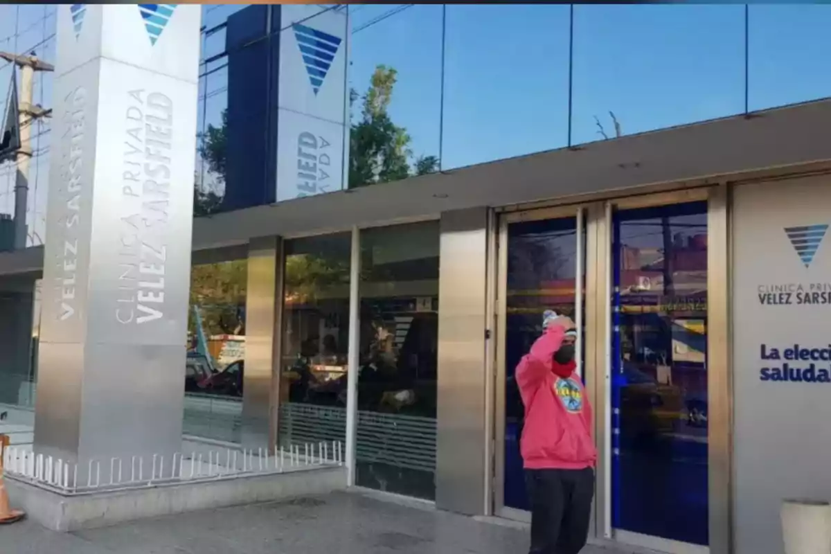 Facade of the Vélez Sarsfield Private Clinic with a person wearing a pink sweatjersey and a face mask standing in front of the main entrance