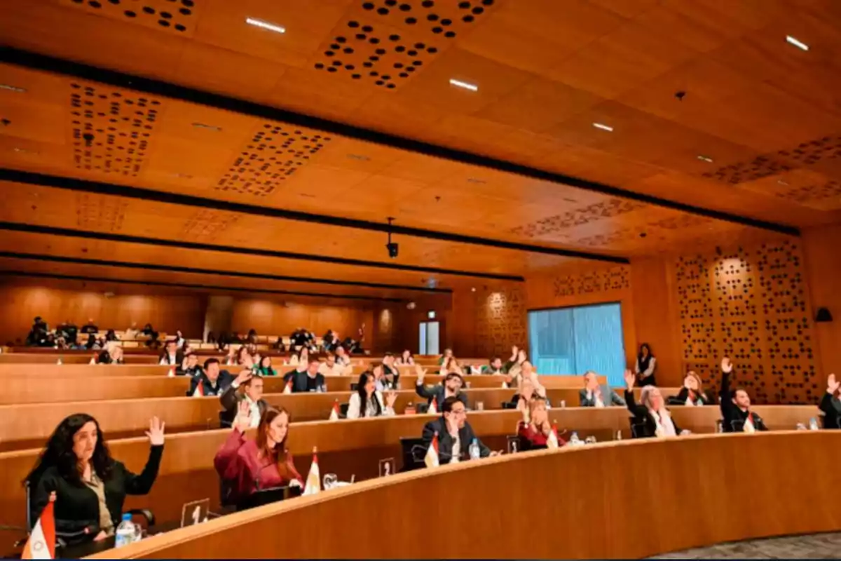 Personas sentadas en un auditorio moderno levantan la mano durante una votación o asamblea Personas sentadas en un auditorio moderno levantan la mano durante una votación o asamblea