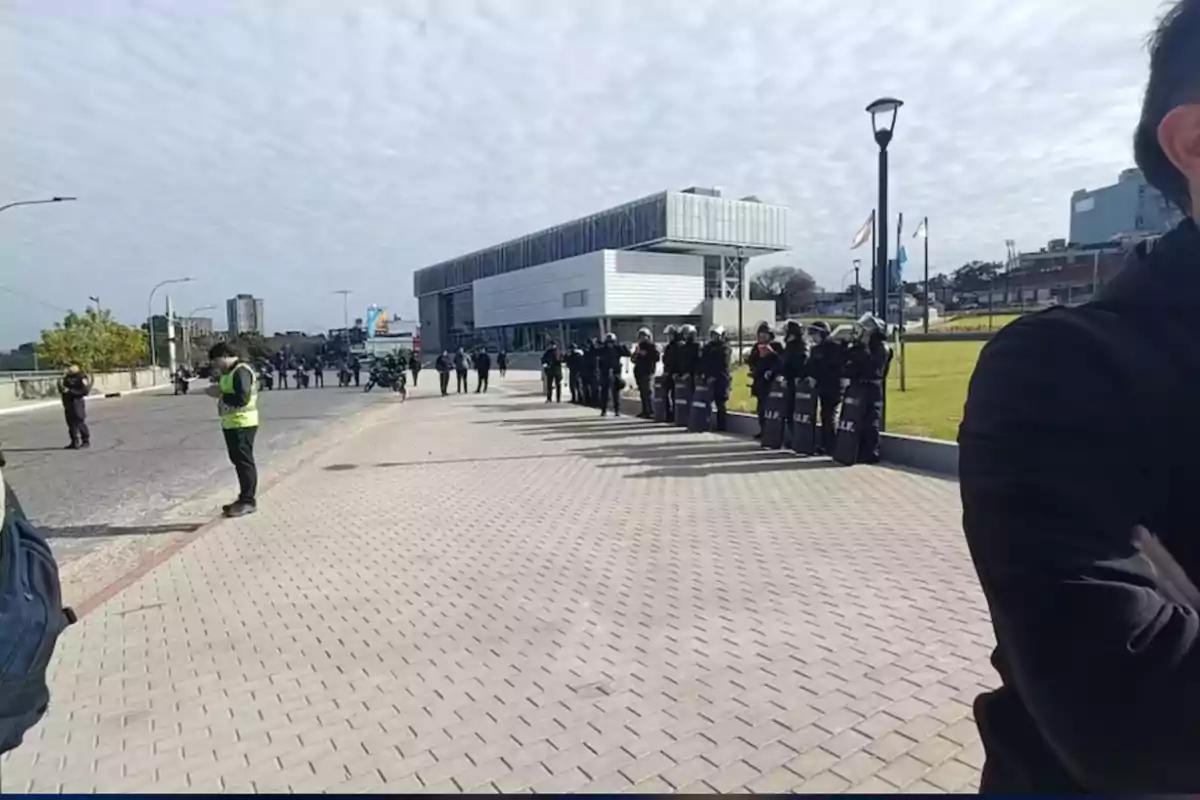 Several police officers with shields form a line in front of a modern building while other people watch and walk around the area.