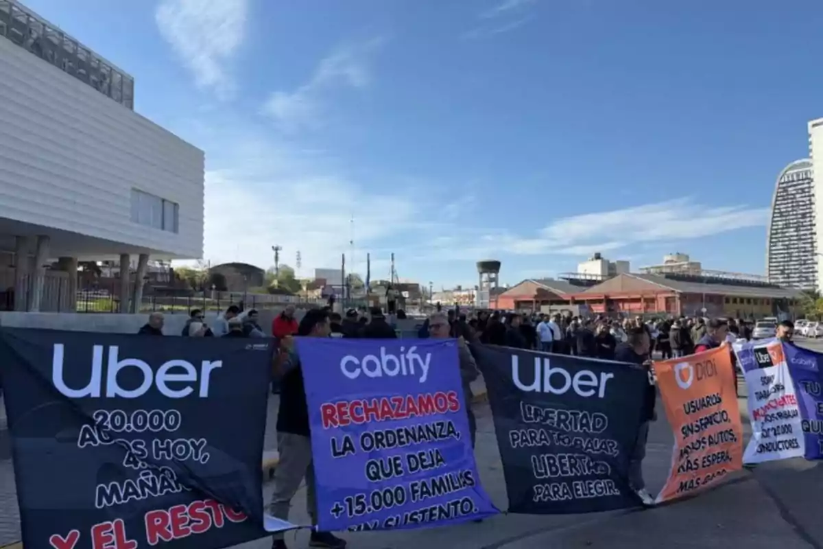 Manifestación de conductores de aplicaciones de transporte como Uber, Cabify y Didi sosteniendo pancartas con mensajes de protesta frente a un edificio moderno y un grupo de personas reunidas en la calle Manifestación de conductores de aplicaciones de transporte como Uber, Cabify y Didi sosteniendo pancartas con mensajes de protesta frente a un edificio moderno y un grupo de personas reunidas en la calle