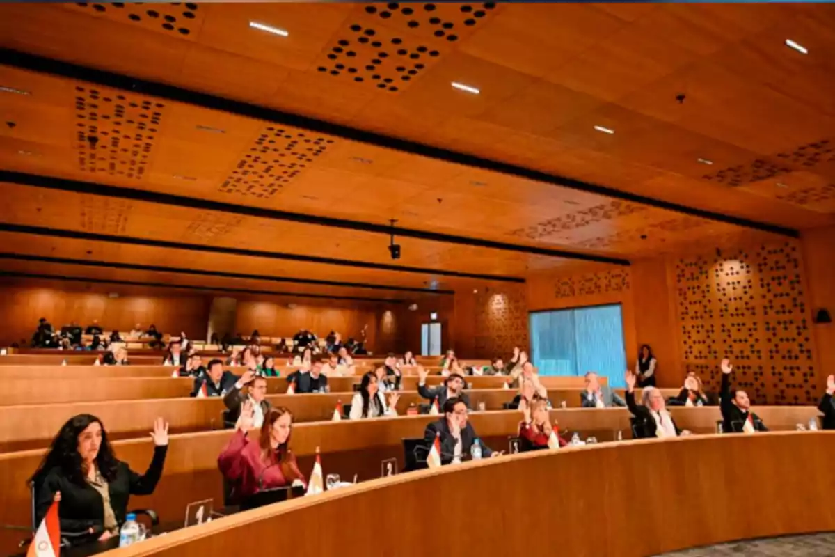 Personas sentadas en un auditorio moderno levantando la mano durante una votación o asamblea Personas sentadas en un auditorio moderno levantando la mano durante una votación o asamblea