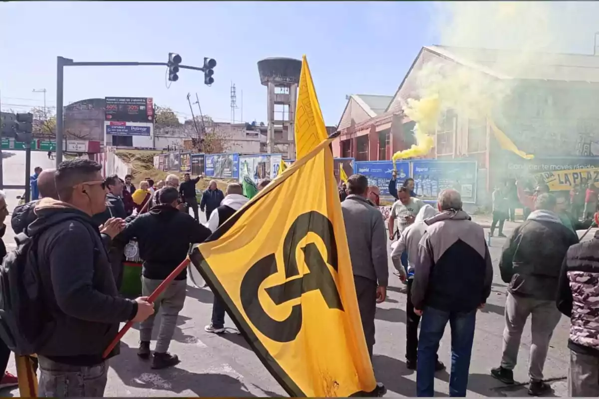 Un grupo de personas participa en una manifestación en la calle, algunas portan banderas amarillas y hay humo amarillo en el ambiente, al fondo se observan edificios industriales y carteles publicitarios.