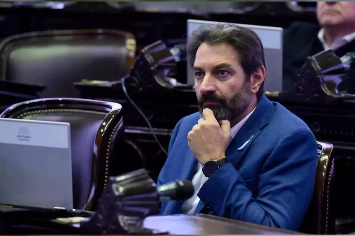 Bearded man in a blue jacket sitting in a parliamentary chamber with a thoughtful expression