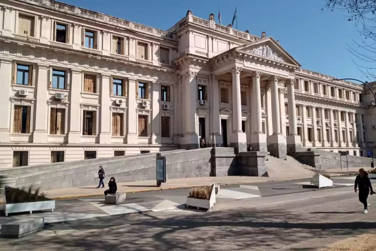 Facade of a historic neoclassical building with columns and staircases, people walking, and clear sky
