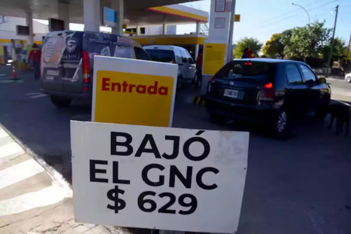 Sign at a gas station announcing the drop in CNG price to 629 pesos while several cars line up to refuel