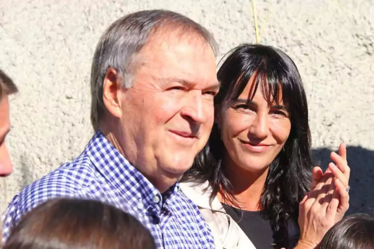 A gray-haired man in a plaid jersey smiles next to a dark-haired woman who is clapping; both are outdoors under the sunlight.