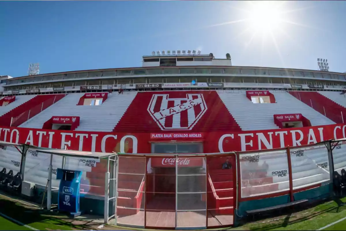 Main stand of a soccer stadium with red and white seats, Instituto Atlético Central Córdoba’s crest in the center, and the sun shining in the sky Main stand of a soccer stadium with red and white seats, Instituto Atlético Central Córdoba’s crest in the center, and the sun shining in the sky