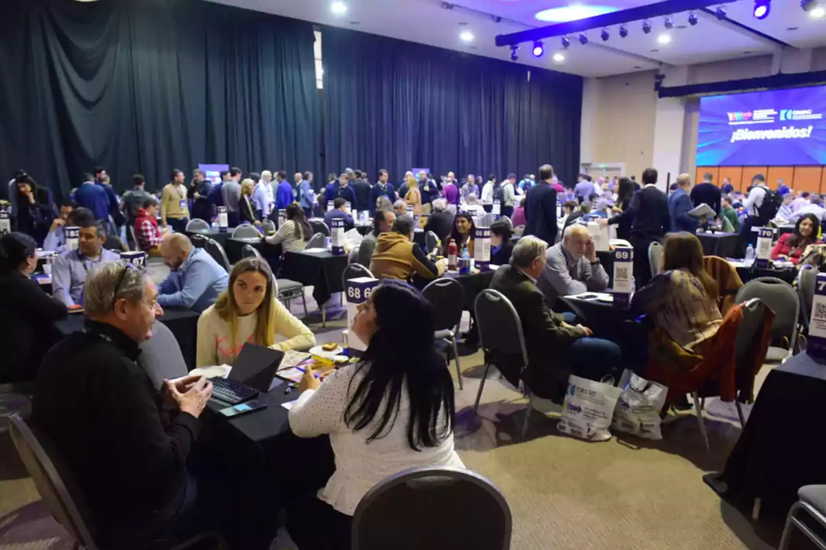 People seated at round tables participating in a business networking event inside a spacious hall with artificial lighting and a large screen in the background displaying a welcome message.