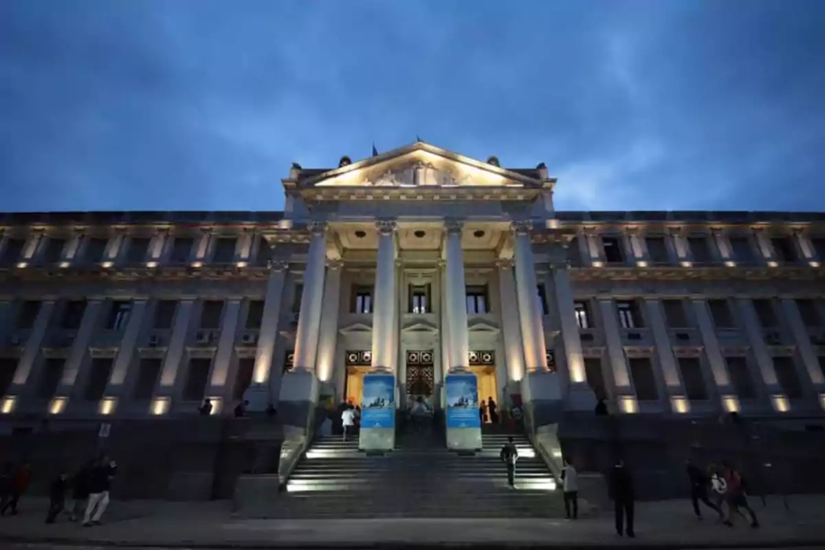 Fachada de un edificio histórico con columnas iluminadas al anochecer y personas subiendo las escaleras principales