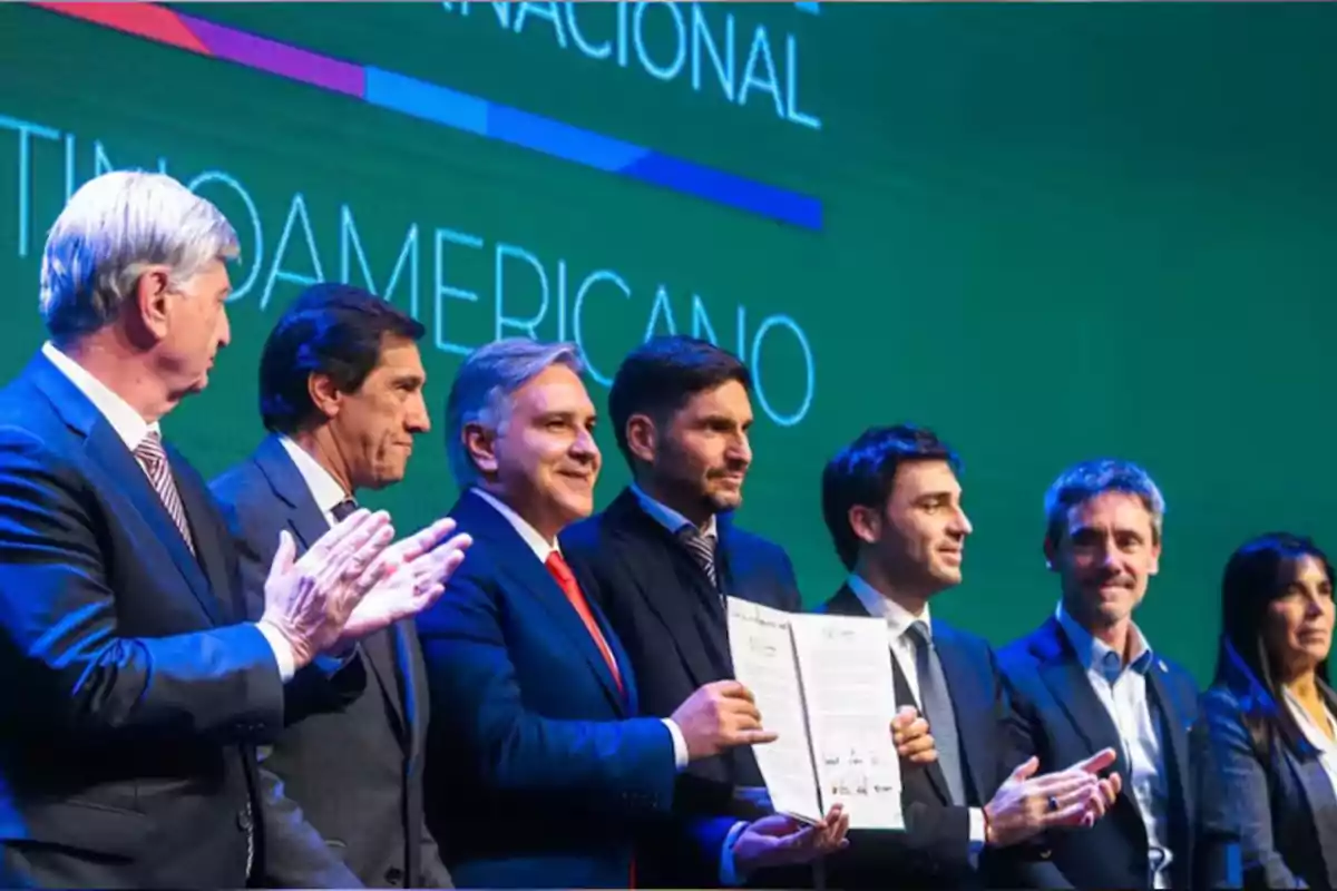 Group of people dressed in suits on a stage during a formal event, one of them holding a document while the others applaud, with a green background and large letters. Group of people dressed in suits on a stage during a formal event, one of them holding a document while the others applaud, with a green background and large letters.