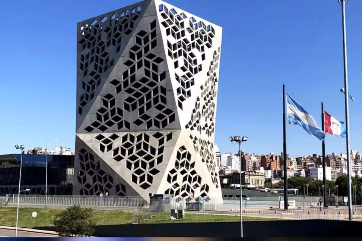 Edificio moderno con diseño geométrico en la fachada, dos banderas ondeando y una ciudad al fondo bajo un cielo despejado Edificio moderno con diseño geométrico en la fachada, dos banderas ondeando y una ciudad al fondo bajo un cielo despejado
