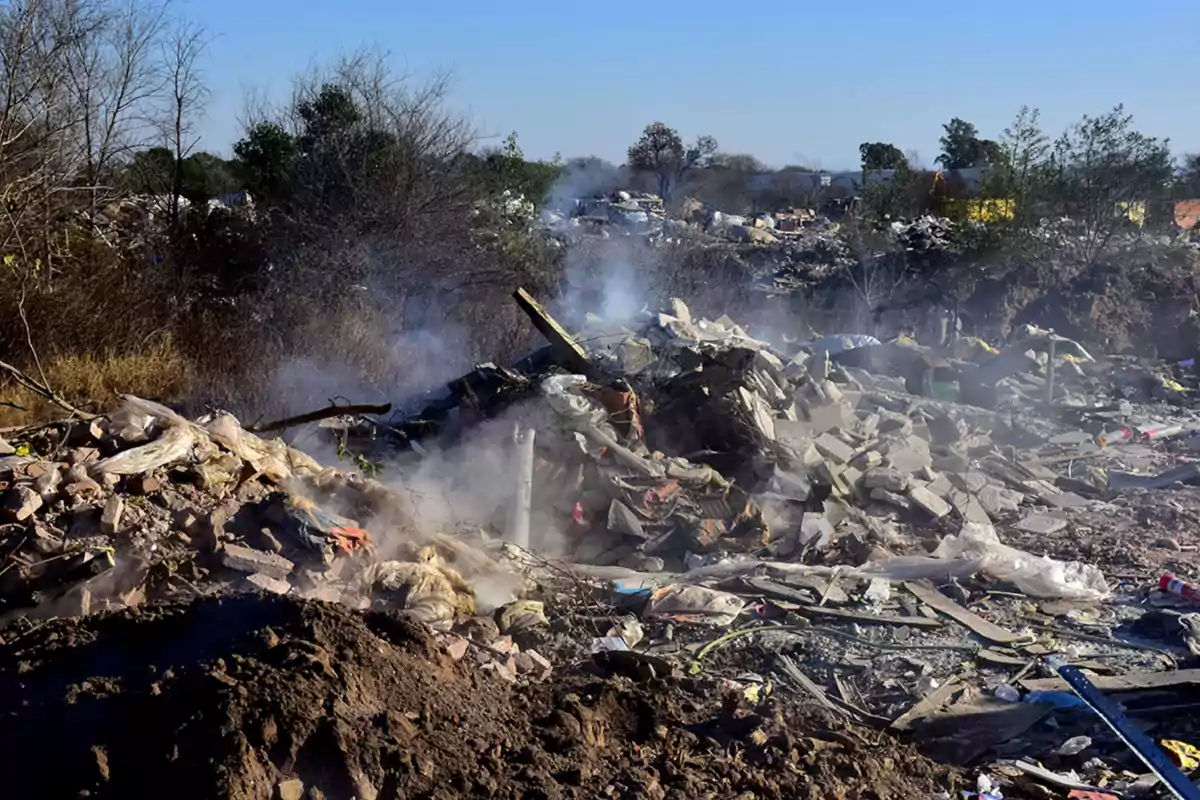 Montón de basura y escombros con humo saliendo entre los desechos en un terreno al aire libre rodeado de árboles secos