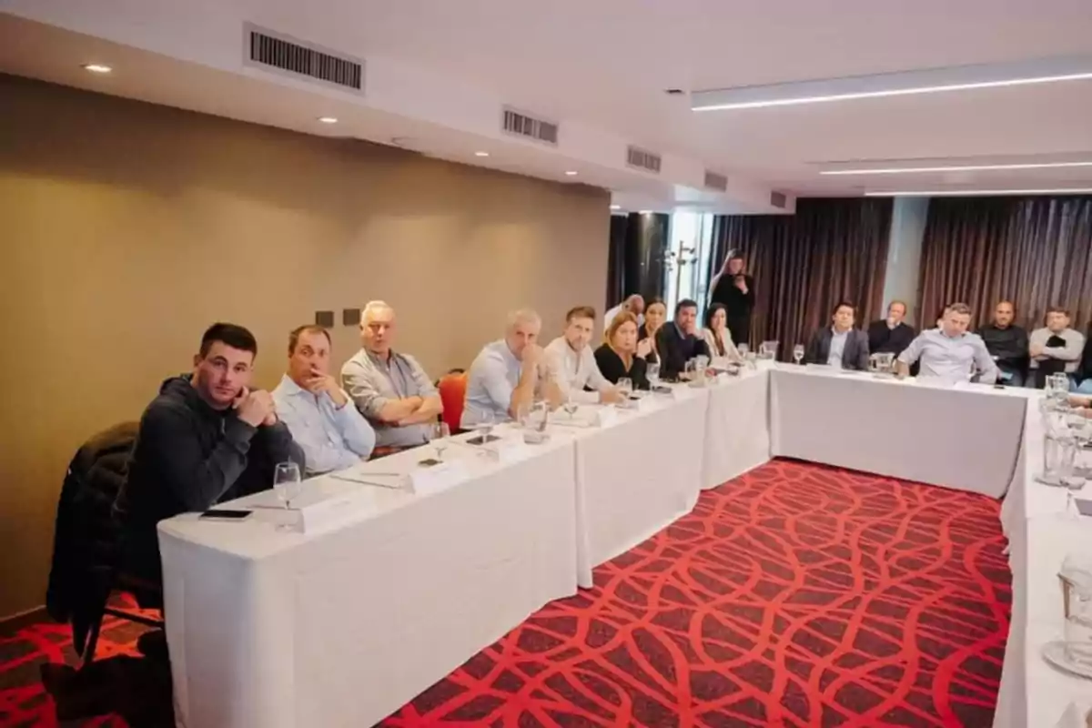 People seated around U-shaped tables in a meeting room, paying attention to a presentation or discussion People seated around U-shaped tables in a meeting room, paying attention to a presentation or discussion