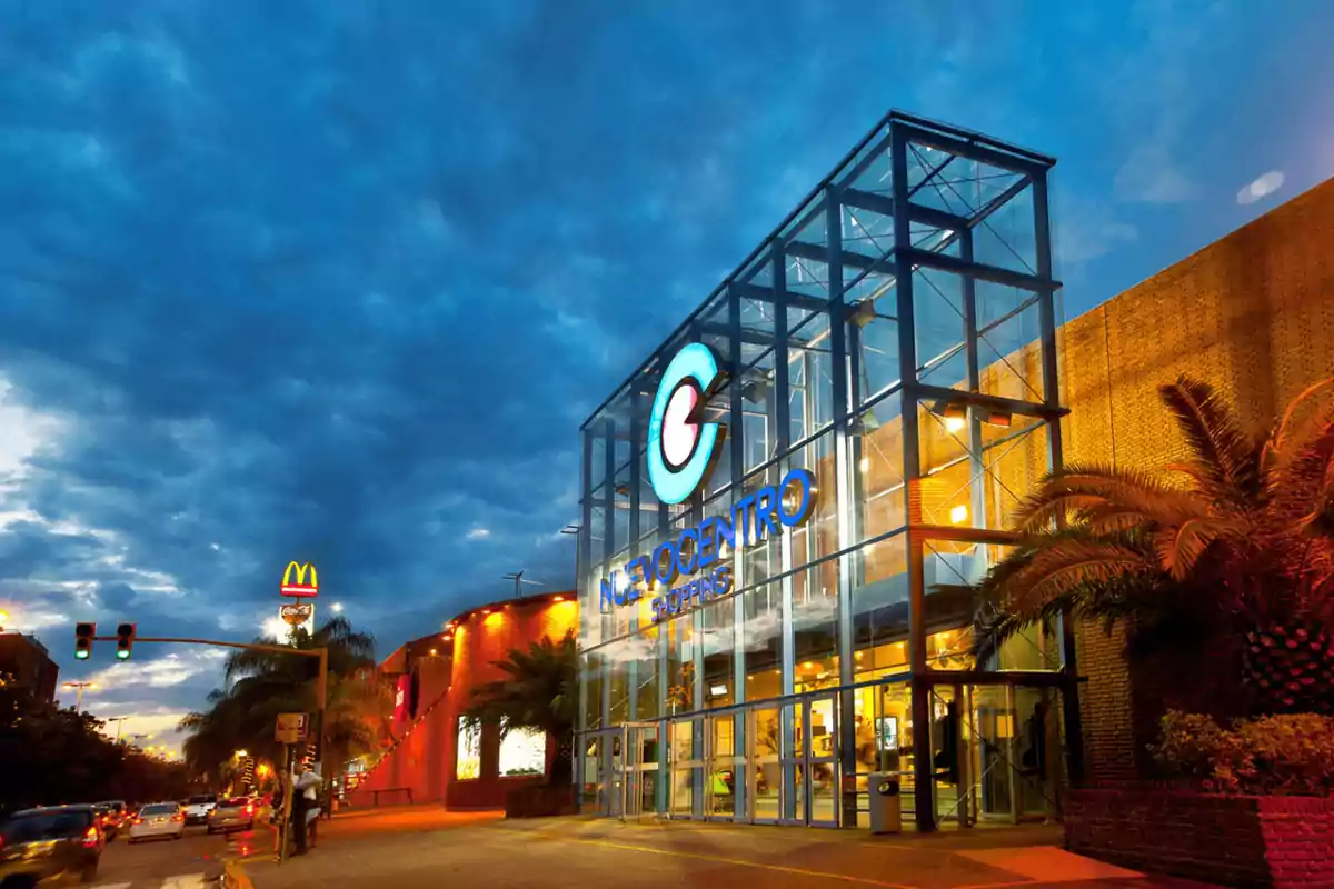 Fachada de un centro comercial moderno con estructura de vidrio iluminada al atardecer y tráfico en la calle