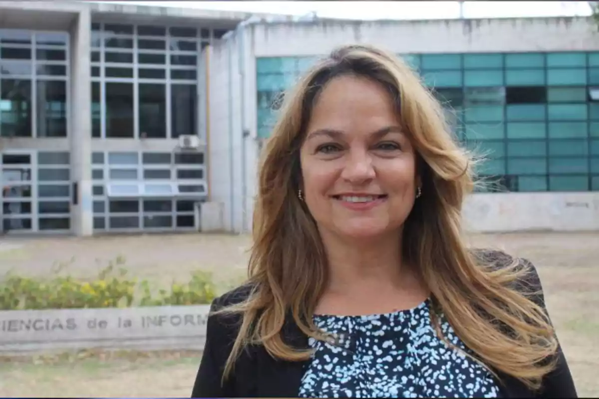 Mujer sonriente de cabello rubio oscuro frente a un edificio moderno con ventanas de vidrio y paredes de concreto